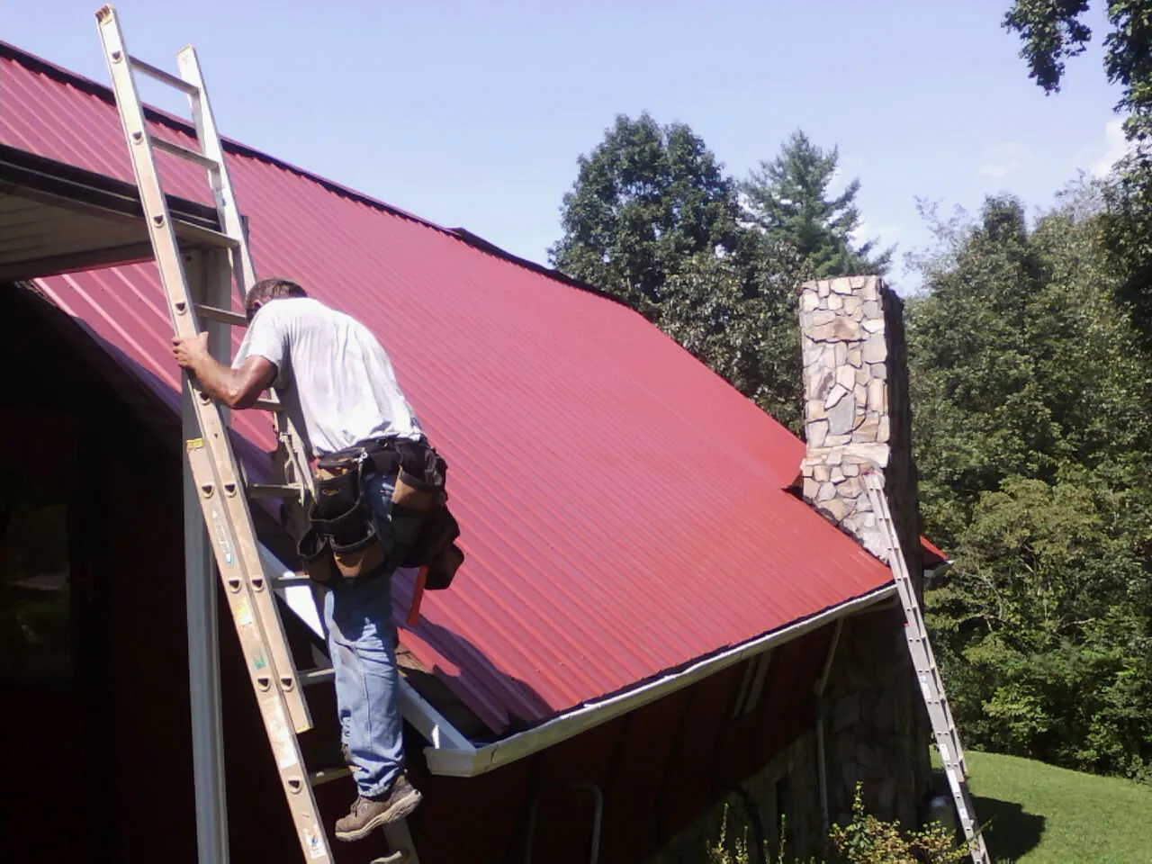 Roofer on a ladder beside a red metal roof and stone chimney — Soffit & Fascia Repair by Roofing Brothers in Lyles